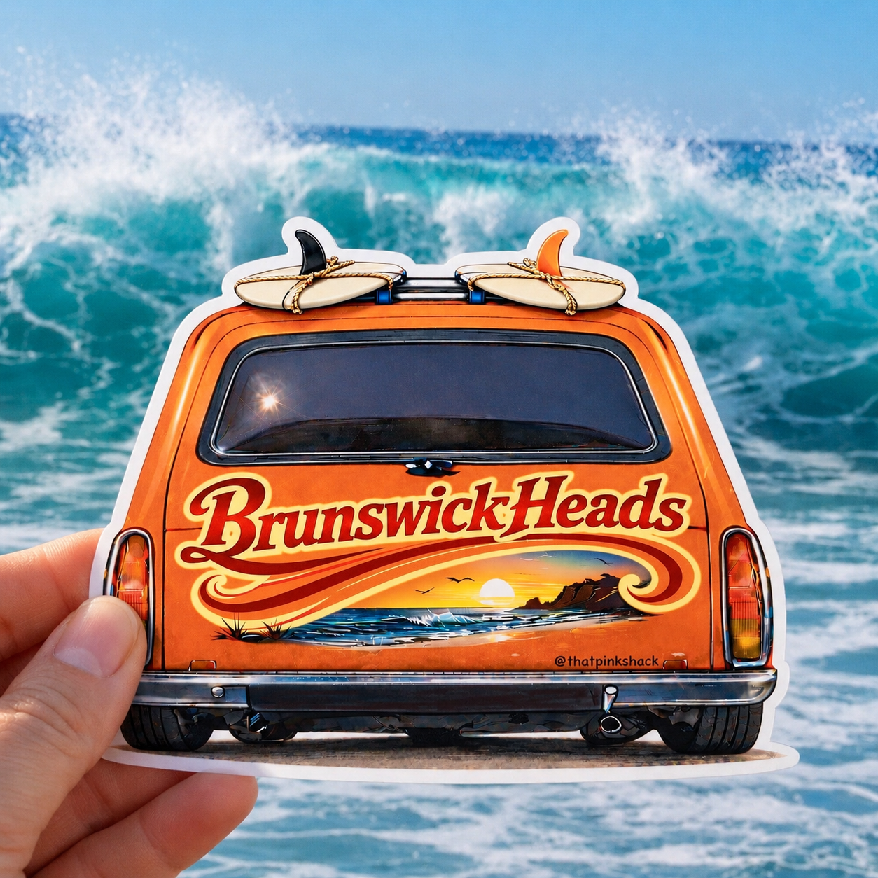 A hand holds a die-cut sticker shaped like the back of an orange station wagon with surfboards on top and a beach sunset graphic. The sticker has the words "Brunswick Heads" in red script.