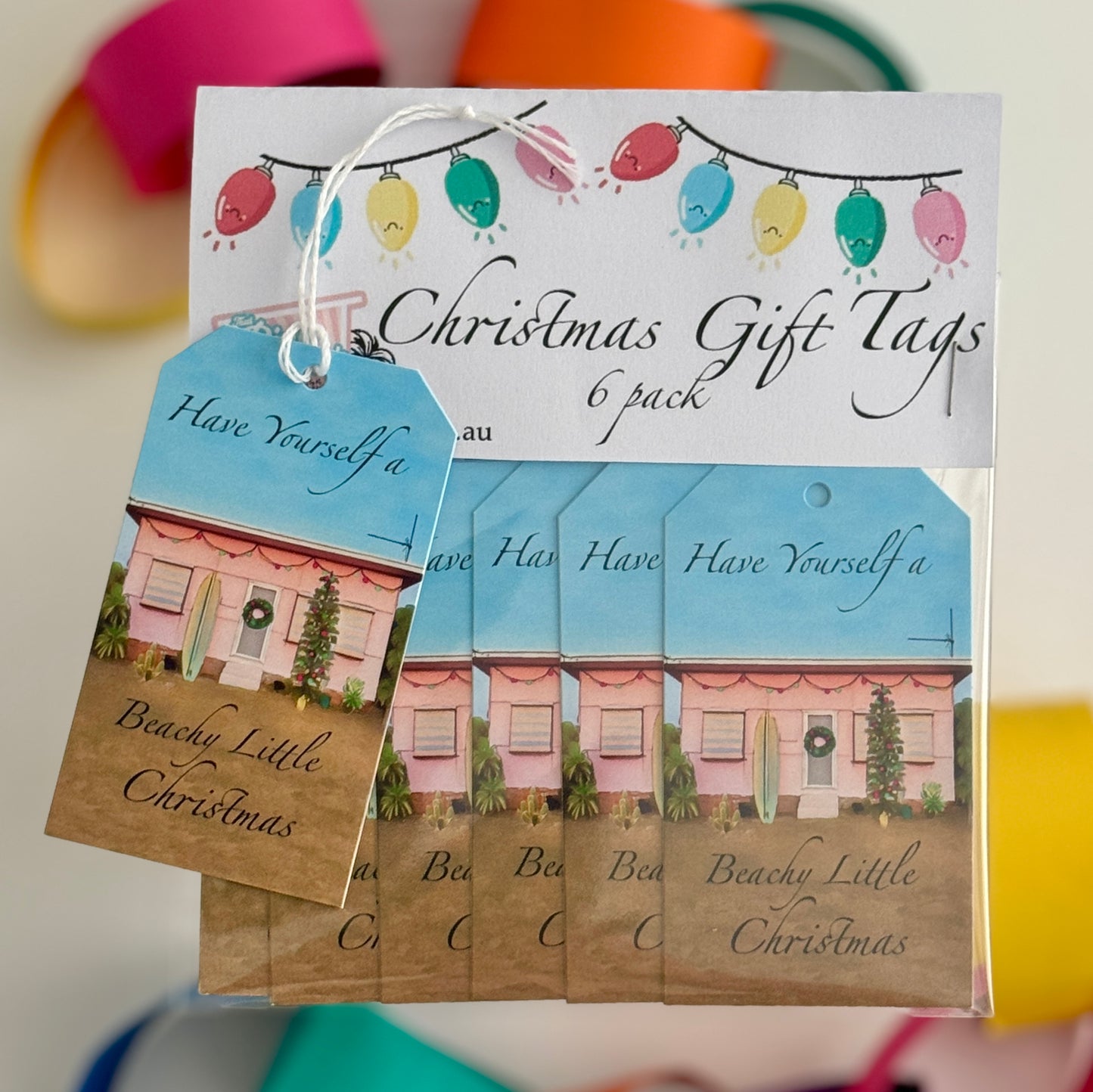 “A packaged 6-pack set of beach-themed 6 Christmas gift tags featuring a retro pink beach shack decorated with lights, a surfboard, and a Christmas tree. One tag is displayed on top with white string attached, showing the phrase ‘have yourself a beach little Christmas !’ Decorative colourful Christmas lights are printed on the header card above.”