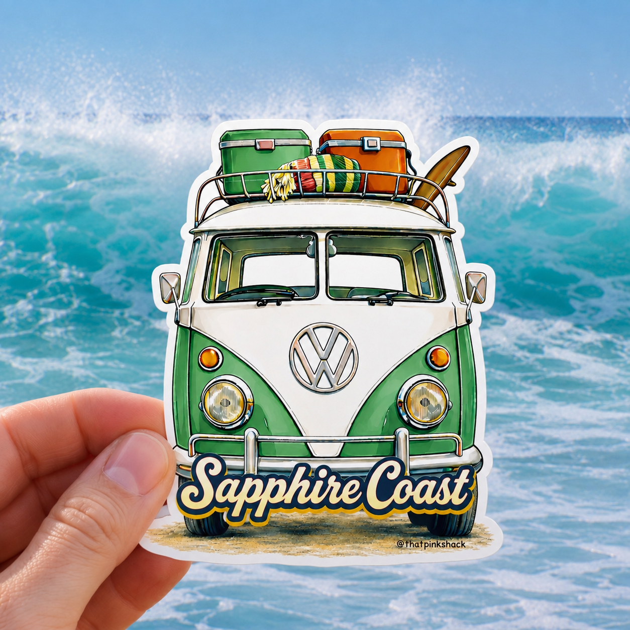 A hand holds a die-cut sticker featuring a retro green and white Volkswagen camper van with luggage and a surfboard on its roof, set against a real ocean wave background. The sticker includes the text 'Sapphire Coast'