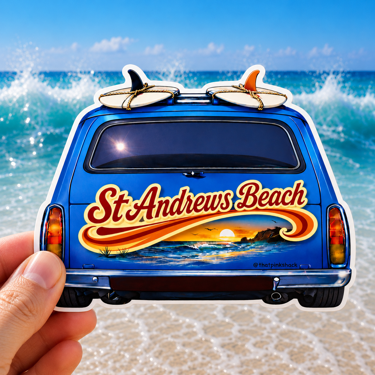 A die-cut sticker shaped like the back of a blue van with surfboards on the roof and a sunset beach graphic, held by a hand against a beach and ocean background. The van has large red lettering reading 'St.Andrews Beach'