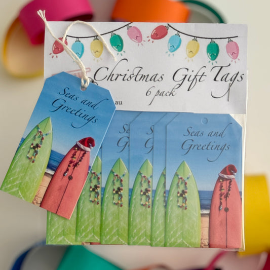 “A packaged 6-pack of beach-themed Christmas gift tags featuring two surfboards decorated with festive lights, one wearing a Santa hat, against a seaside background. One tag with a white string is displayed on top, showing the phrase ‘Seas and Greetings.’ The header card above includes colourful Christmas light illustrations.”