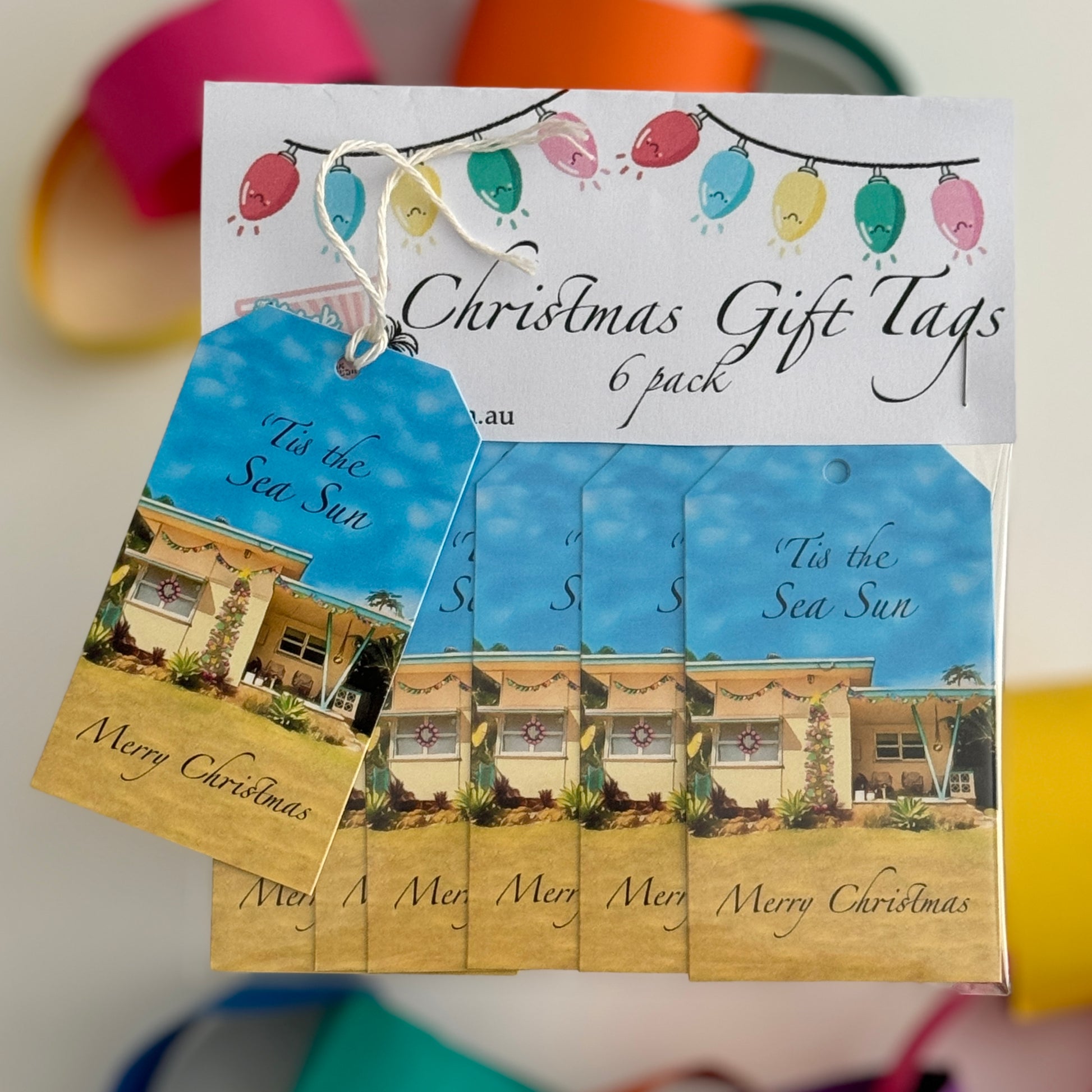 “A packaged 6-pack set of beach-themed 6 Christmas gift tags featuring a retro yellow beach shack decorated with lights and a Christmas tree . One tag is displayed on top with white string attached, showing the phrase ‘Deck the Palms!’ Decorative colourful Christmas lights are printed on the header card above.”
