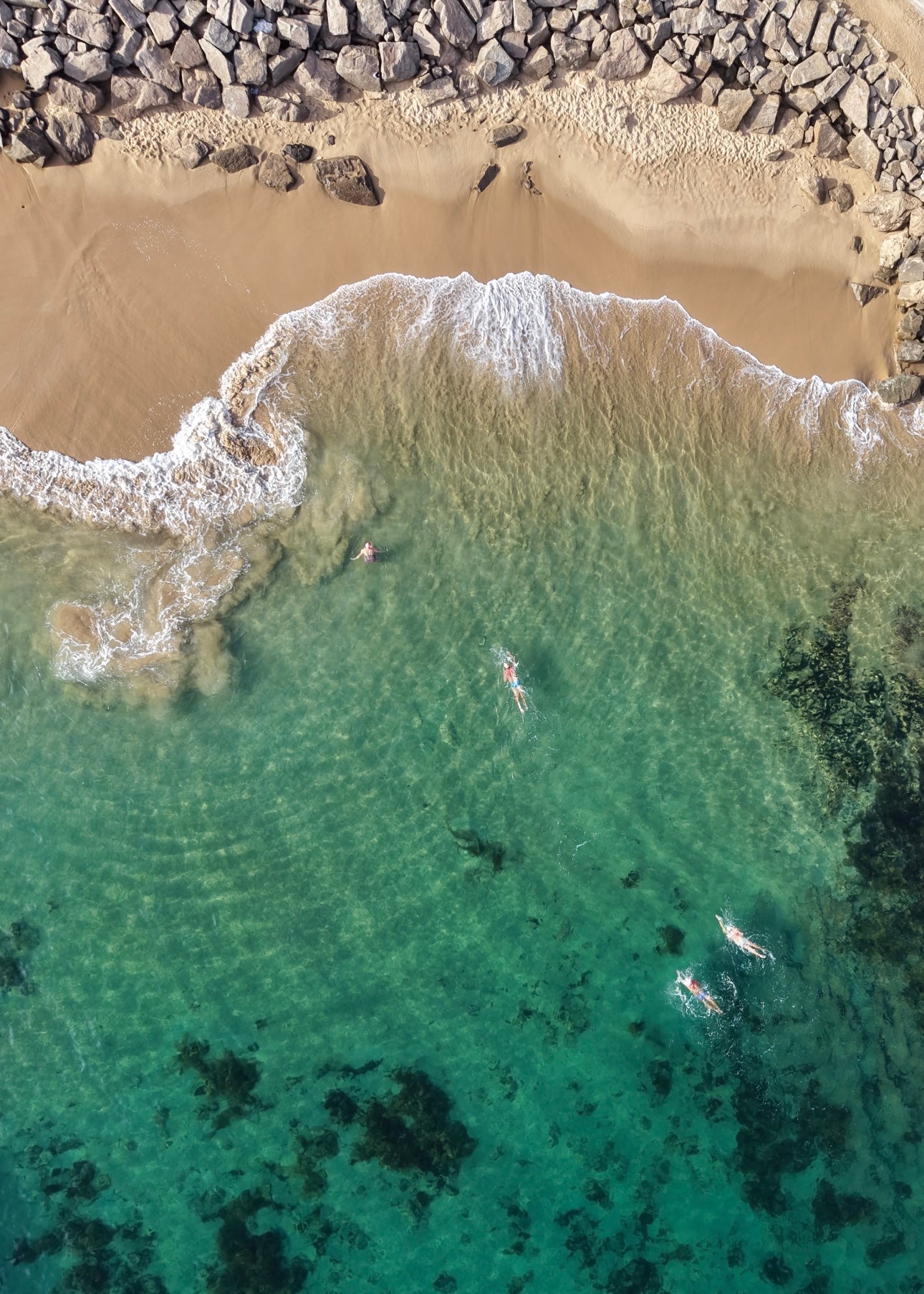 Aerial view of people swimming in clear turquoise water with a sandy beach and rocks.