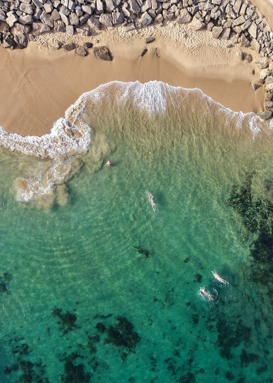 Aerial view of people swimming in clear turquoise water with a sandy beach and rocks.