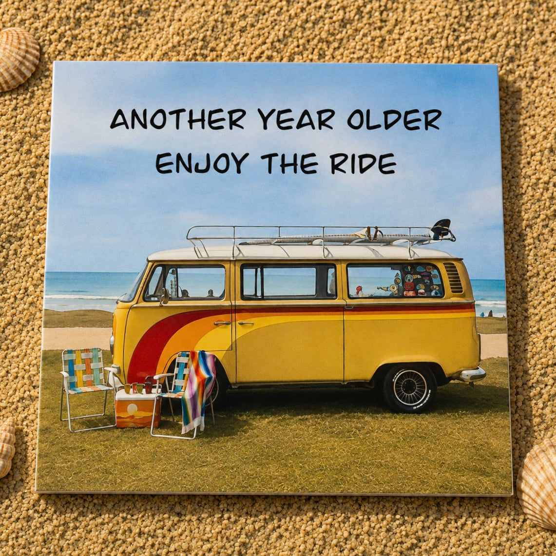 A square greeting card featuring a retro yellow and red VW bus on a beach with the text 'ANOTHER YEAR OLDER ENJOY THE RIDE', displayed on a sandy surface with small seashells