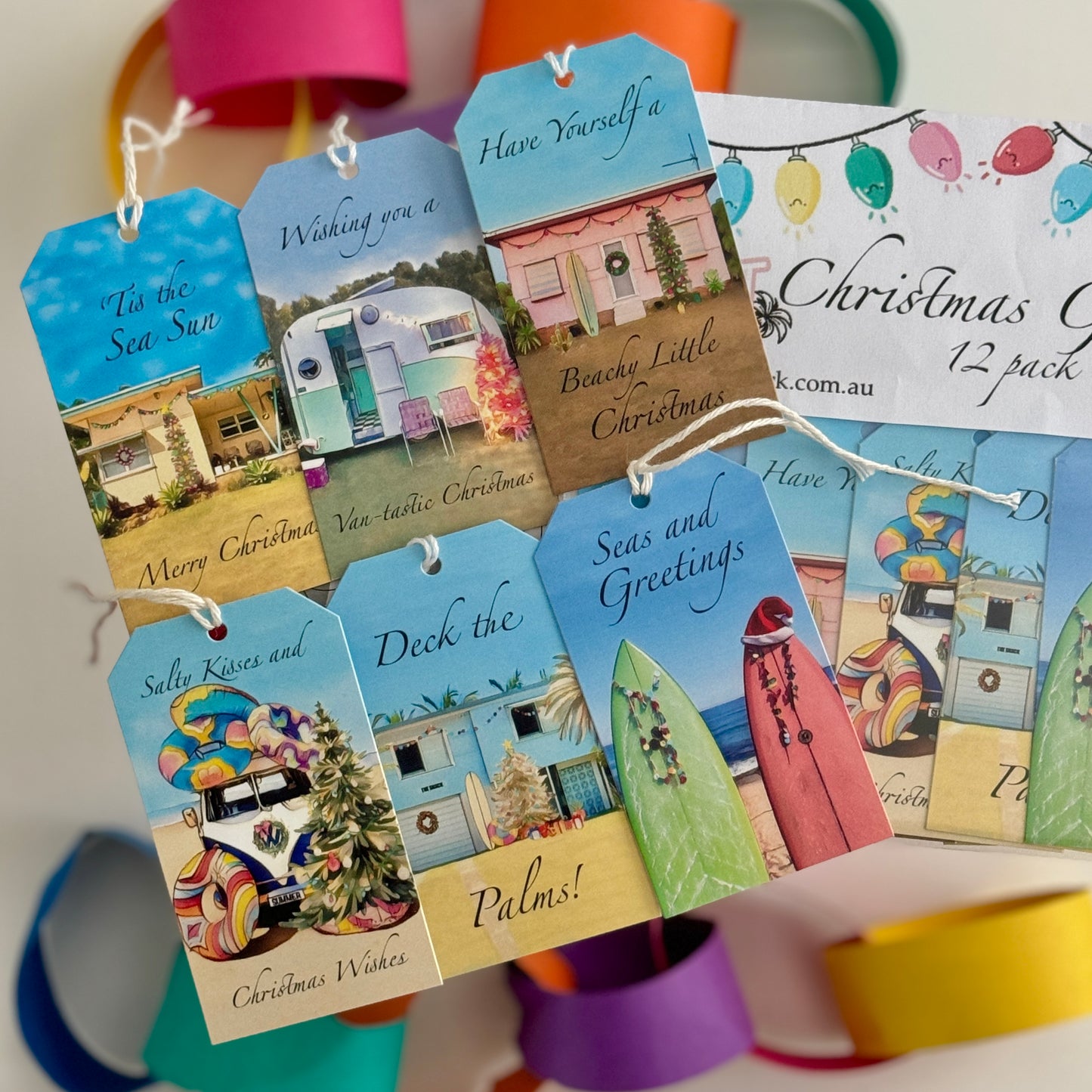 “A set of 12 colourful Aussie beach-themed Christmas gift tags displayed in a fan layout. Each tag features a retro coastal caravan, surfboards, or a festive kombi van, along with playful holiday sayings like ‘Deck the Palms,’ ‘Seas and Greetings,’ and ‘Have Yourself a Beachy Little Christmas.’ The tags are tied with white string and arranged beside a small card showing they come in a 6-pack.”
