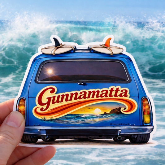 A hand holds a die-cut sticker shaped like the back of a blue van with surfboards on top and a large 'Gunnamatta' logo across the back, set against a background of ocean waves.