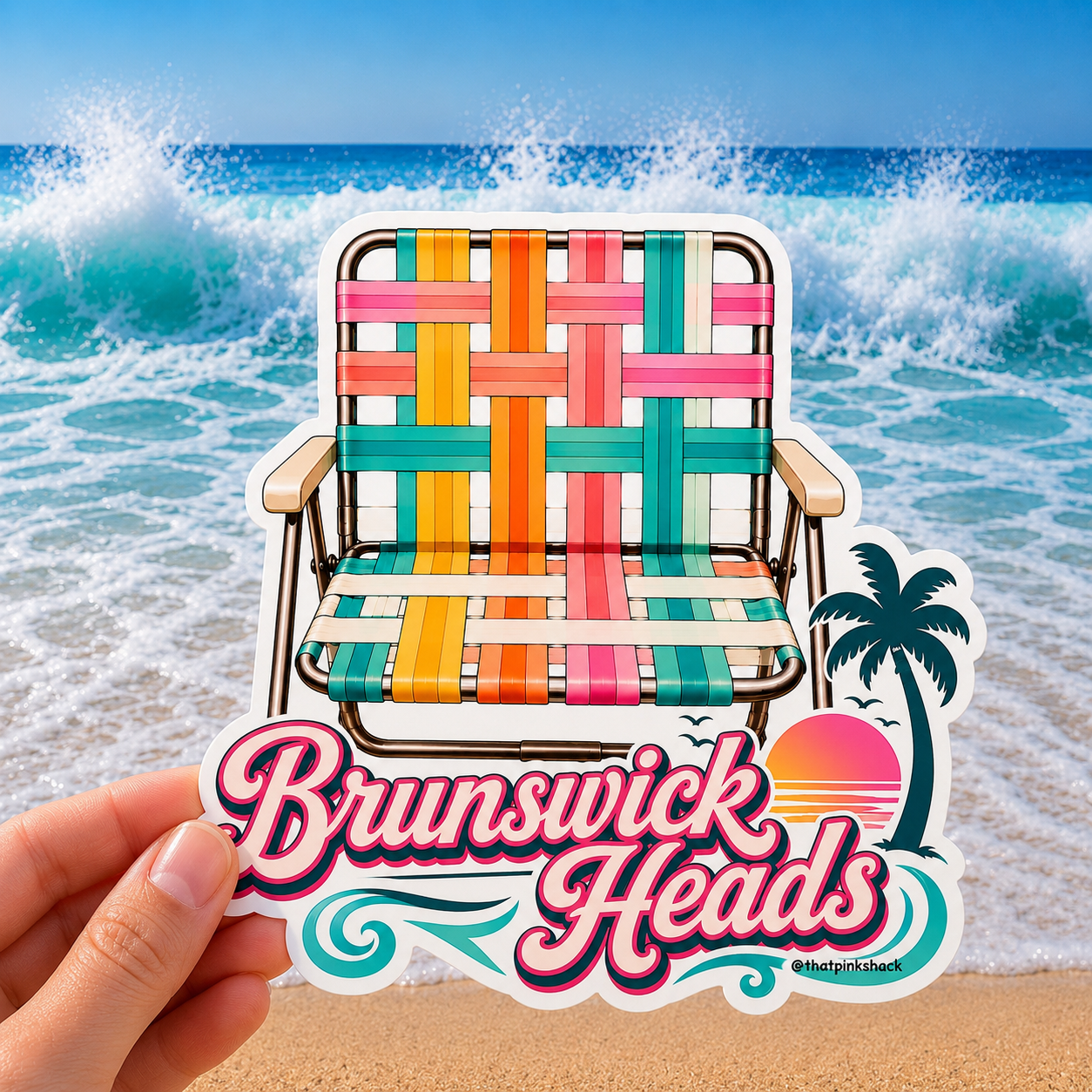 A hand holds a die-cut sticker featuring a colorful retro beach chair and the text 'Brunswick Heads' with a sunset and palm tree, set against a background of ocean waves and sand.