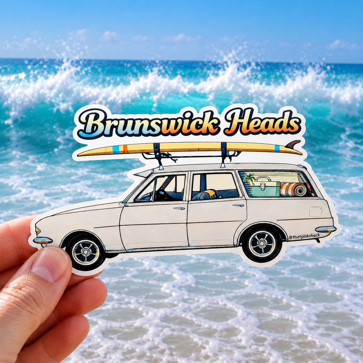 A hand holds a die-cut sticker featuring a vintage white holden station wagon with a surfboard on its roof, set against a beach scene with ocean waves and the text 'Brunswick Heads'.