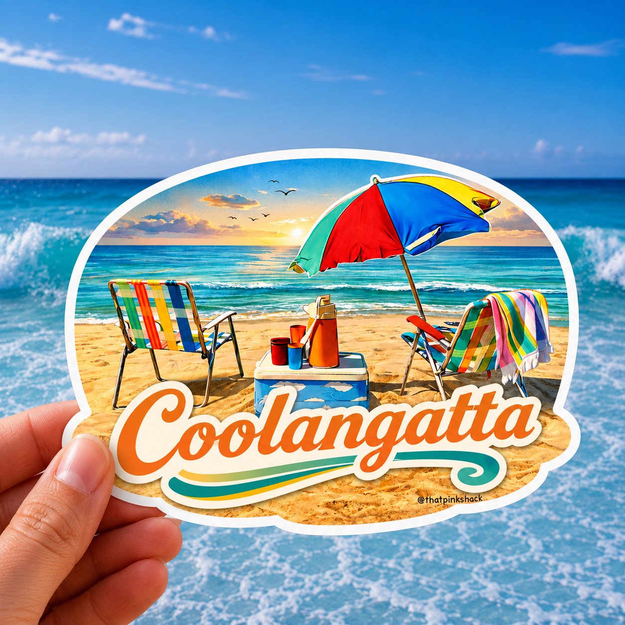 A hand holds an oval sticker featuring a beach scene with two thatched retro camp chairs, a red-blue-yellow umbrella, a vintage esky cooler, and the word "Coolangatta" in orange script. The background shows blue ocean water and sky.