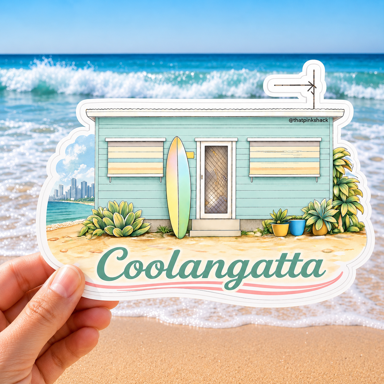 A hand holds a die-cut sticker featuring an illustrated beach house with a surfboard, plants, and the text 'Coolangatta' against a real beach and ocean background.