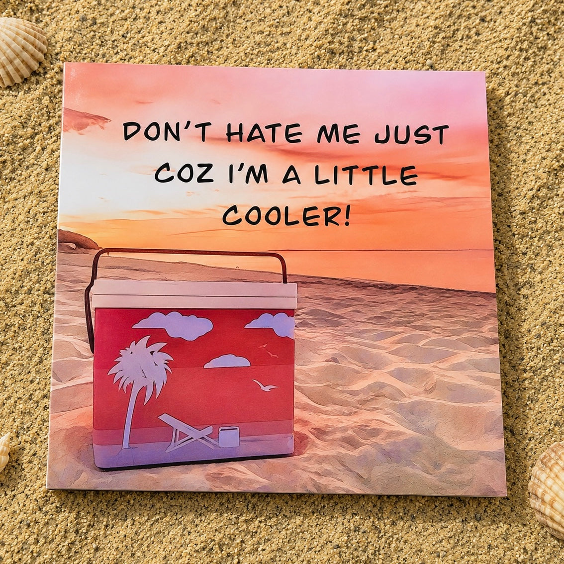 Square greeting card with don’t hate me just coz I’m a little cooler written on it laying flat on the sand 