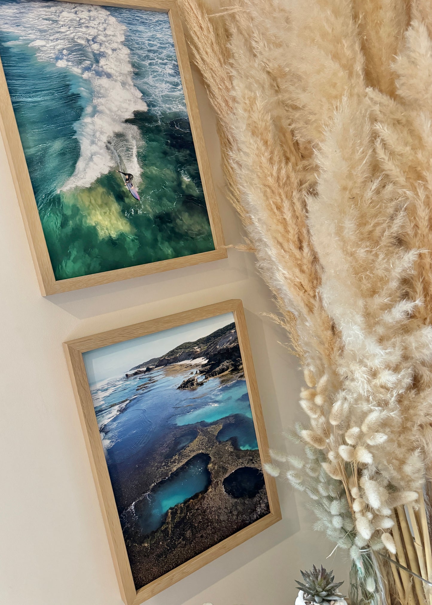 Two framed artworks of a surfer in a reef break and the rock pools of on a wall with pampas grass.