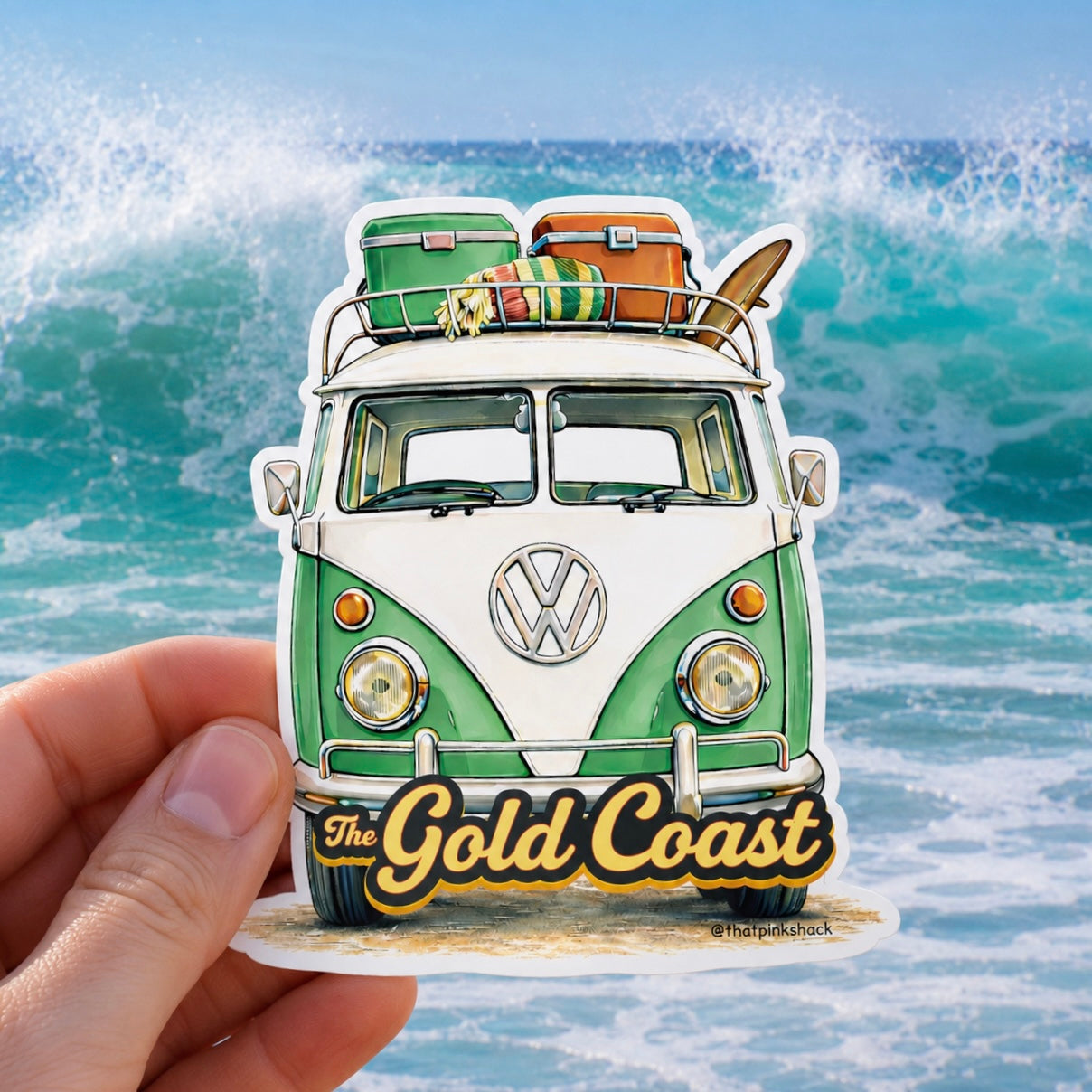A hand holds a die-cut sticker featuring a vintage green and white Volkswagen bus with luggage and a surfboard on its roof, set against a background of ocean waves. The sticker includes the text 'The Gold Coast' in yellow script.