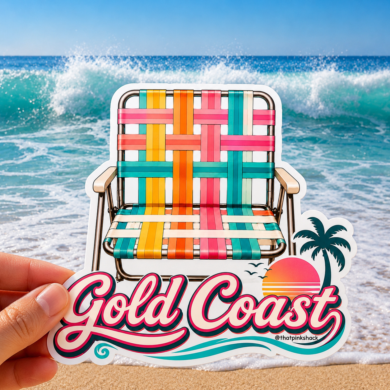 A hand holds a die-cut sticker featuring a colorful retro beach chair in front of a real ocean wave scene. The sticker has the words "Gold Coast" in large pink script with red and blue outlines, plus a small palm tree and sunset graphic.