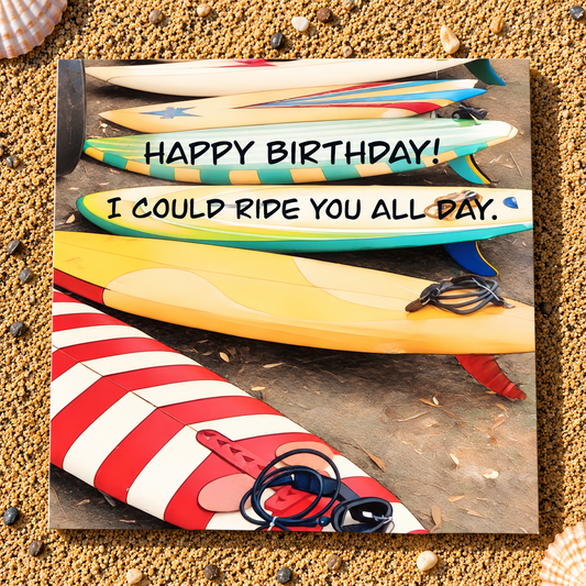 A square printed greeting card featuring a colorful graphic of stacked surfboards with the text 'HAPPY BIRTHDAY! I COULD RIDE YOU ALL DAY.' placed on a sandy beach background with seashells.