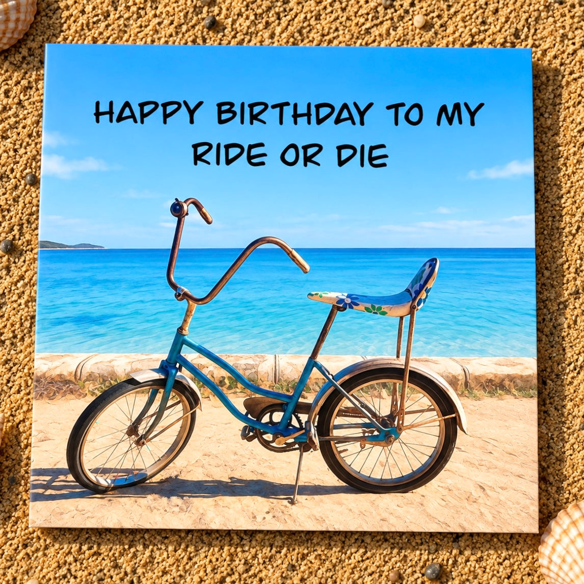 A square printed card featuring a beach scene with a blue cruiser bicycle and the text 'HAPPY BIRTHDAY TO MY RIDE OR DIE,' placed on a sandy textured surface.