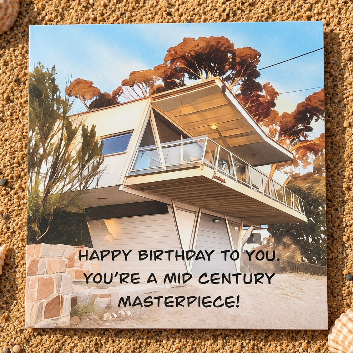 A square greeting card  featuring a modern mid-century style house with the text 'HAPPY BIRTHDAY TO YOU. YOU'RE A MID CENTURY MASTERPIECE!' placed on a sandy surface.