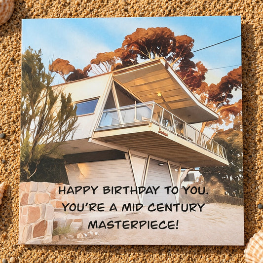 A square greeting card  featuring a modern mid-century style house with the text 'HAPPY BIRTHDAY TO YOU. YOU'RE A MID CENTURY MASTERPIECE!' placed on a sandy surface.