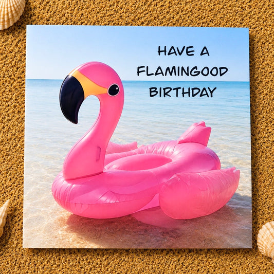 A square printed card featuring a pink flamingo float on a beach with the text 'HAVE A FLAMINGOOD BIRTHDAY,' photographed on a sandy surface with small seashell props around it.