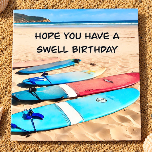 A square greeting card featuring a beach scene with surfboards on sand and the text 'HOPE YOU HAVE A SWELL BIRTHDAY', placed on a sandy surface.