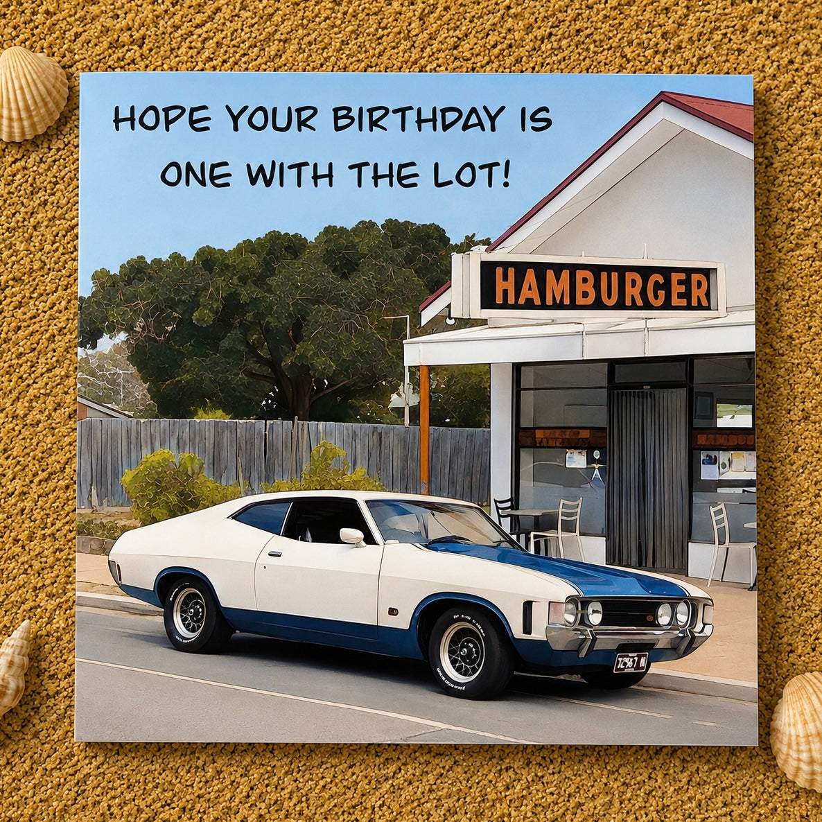 A square birthday card  featuring a vintage white and blue muscle car parked in front of a small hamburger restaurant, with the text 'HOPE YOUR BIRTHDAY IS ONE WITH THE LOT!' at the top. The card is centered on a textured sand background with small shell decorations in the corners.