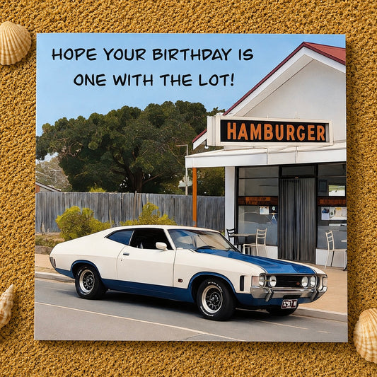 A square birthday card  featuring a vintage white and blue muscle car parked in front of a small hamburger restaurant, with the text 'HOPE YOUR BIRTHDAY IS ONE WITH THE LOT!' at the top. The card is centered on a textured sand background with small shell decorations in the corners.