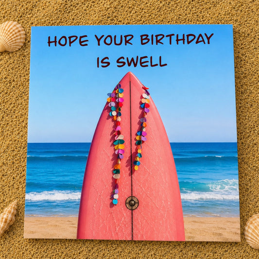 A square greeting card featuring a beach scene with a pink surfboard adorned with colorful heart-shaped decorations, with the text 'HOPE YOUR BIRTHDAY IS SWELL' at the top. The card is displayed on a sandy surface with seashells around it.