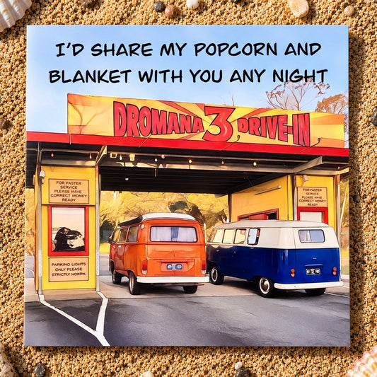 A square printed greeting card featuring a stylized scene from 'Dromana 3 Drive-In' with two vintage vw buses outside theatre, plus the text 'I'D SHARE MY POPCORN AND BLANKET WITH YOU ANY NIGHT.' The print is photographed lying on a sandy textured surface.
