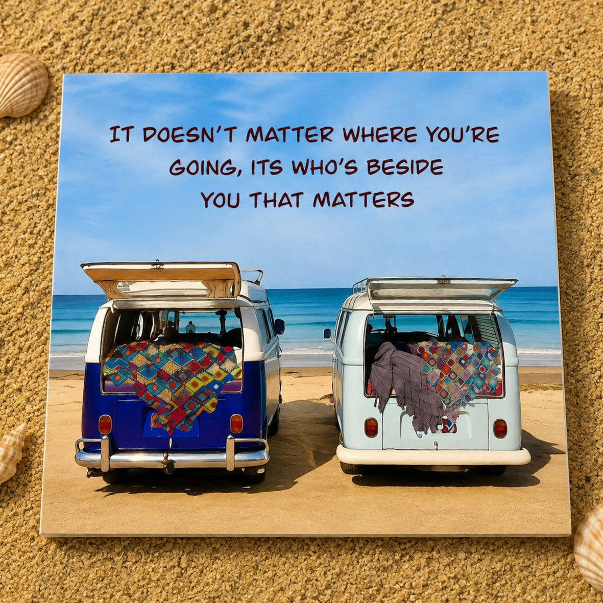 A square greeting card featuring a beach scene with two vans parked on sand, each draped with a colorful patchwork quilt, under a blue sky with the text 'IT DOESN'T MATTER WHERE YOU'RE GOING, ITS WHO'S BESIDE YOU THAT MATTERS'. The card is displayed on a sandy surface with small seashells at the corners.