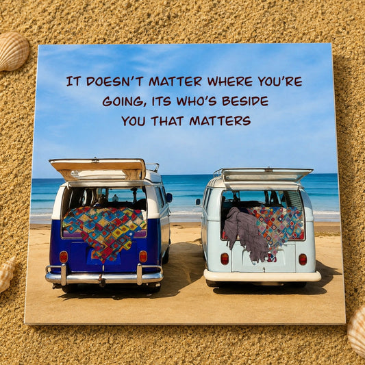 A square greeting card featuring a beach scene with two vans parked on sand, each draped with a colorful patchwork quilt, under a blue sky with the text 'IT DOESN'T MATTER WHERE YOU'RE GOING, ITS WHO'S BESIDE YOU THAT MATTERS'. The card is displayed on a sandy surface with small seashells at the corners.