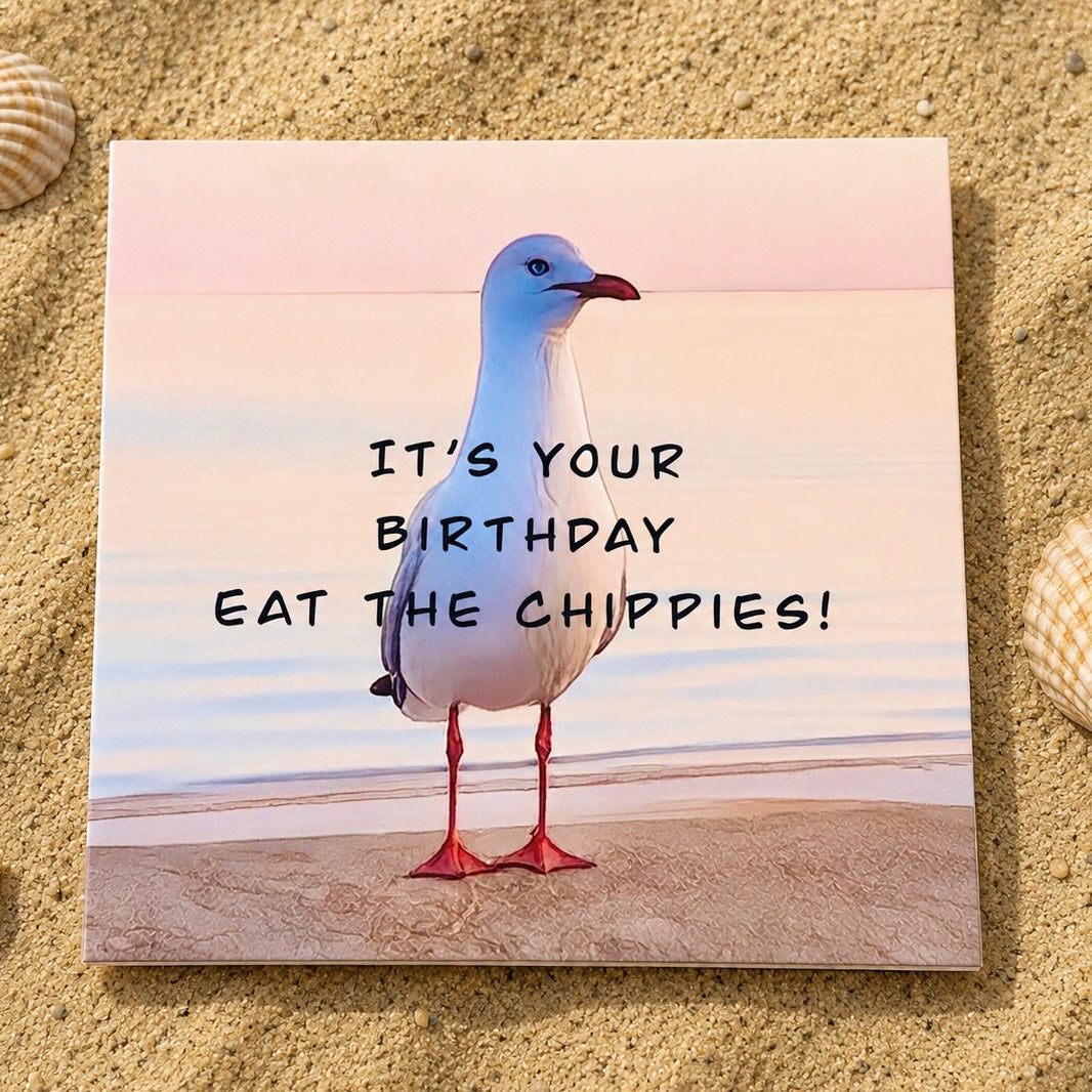 A square printed greeting card  featuring a seagull standing on a beach with the text 'IT'S YOUR BIRTHDAY EAT THE CHIPPIES!' placed on sand between two seashells.