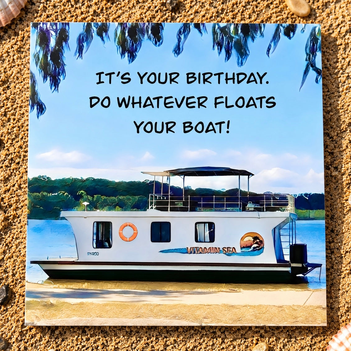 A square printed card featuring a houseboat scene with the text 'IT'S YOUR BIRTHDAY. DO WHATEVER FLOATS YOUR BOAT!' placed on a sandy textured surface.