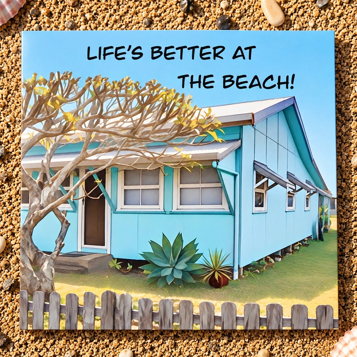 A square printed greeting card featuring an illustrated light blue beach house, green grass, a tree, and the text “LIFE’S BETTER AT THE BEACH!” on a sandy background.