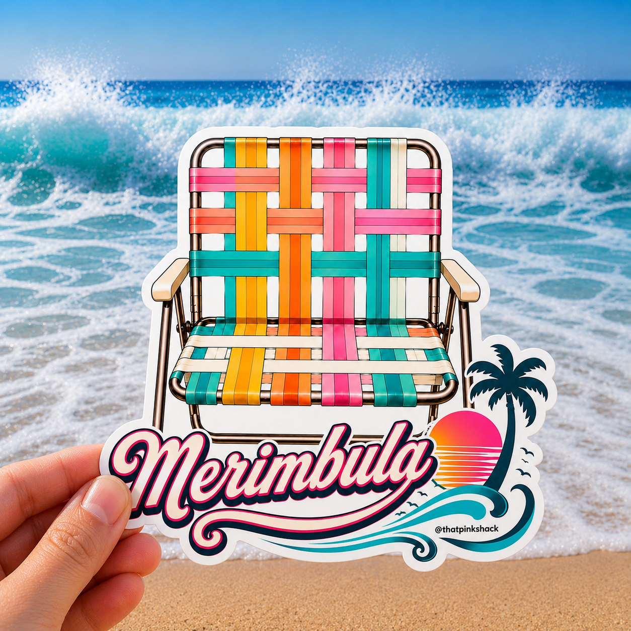 A hand holds a die-cut sticker in front of a beach scene with ocean waves and sand. The sticker shows a colorful woven retro beach chair and the text “Merimbula” with a sunset and palm tree graphic.