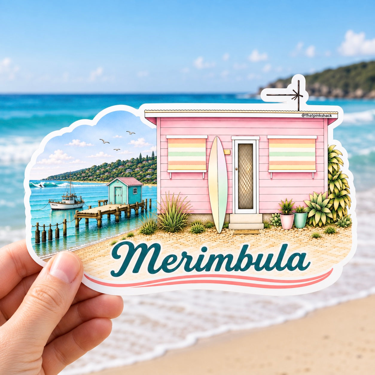 A hand holds a die-cut sticker featuring a pastel beach hut scene with the text 'Merimbula' and a small sign reading 'Merimbula Beach'. The background shows a blurred beach and ocean with a boat.