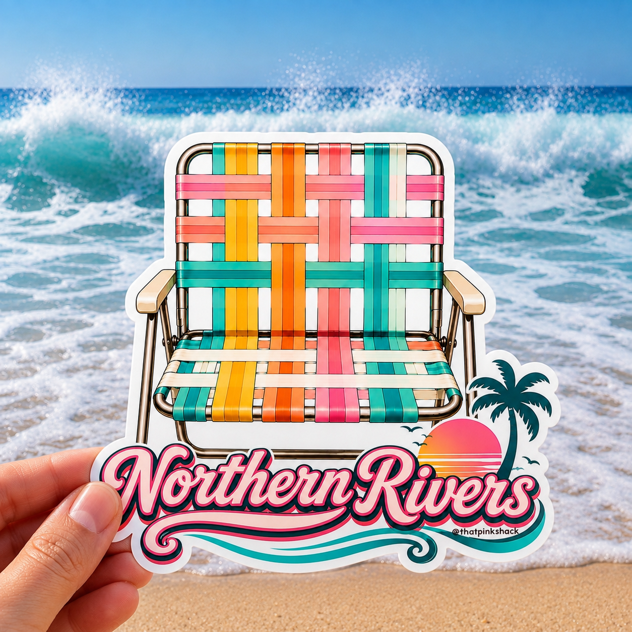 A hand holds a die-cut sticker featuring a colorful woven retro beach chair and the text 'Northern Rivers' with a sunset and palm tree, set against a beach background with ocean waves.