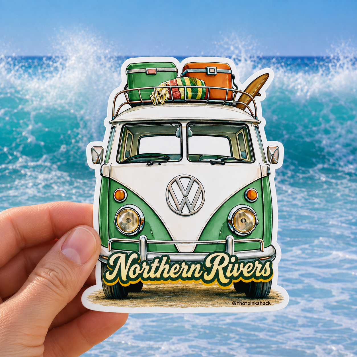 A hand holds a die-cut sticker featuring a green and white vintage Volkswagen camper van with surfboards and coolers on top, set against a background of ocean waves. The front of the van reads 'Northern Rivers'.
