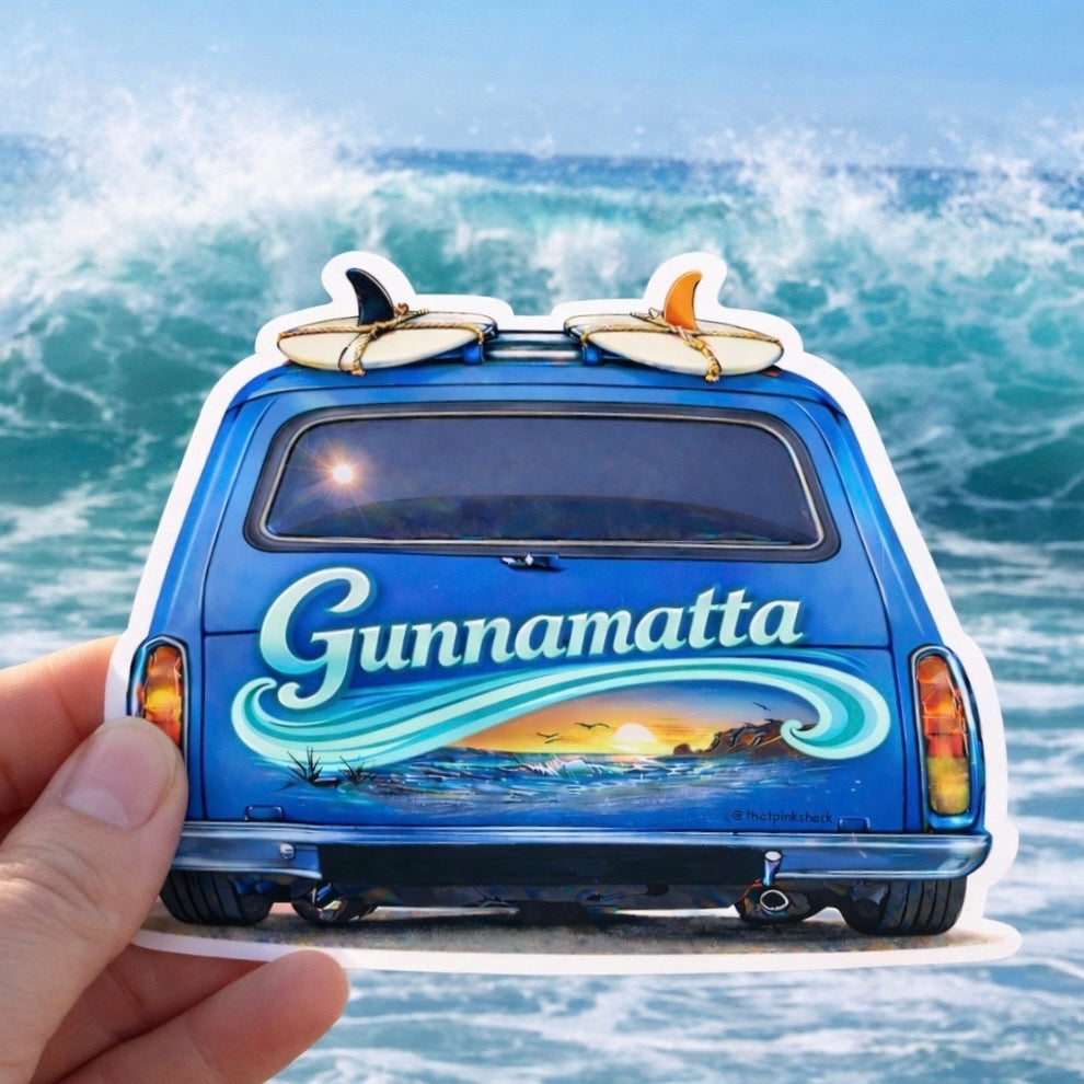 A hand holds a die-cut sticker shaped like the back of a blue van with surfboards on top and the word "Gunnamatta" in large white script over an ocean sunset illustration. The background shows ocean waves.