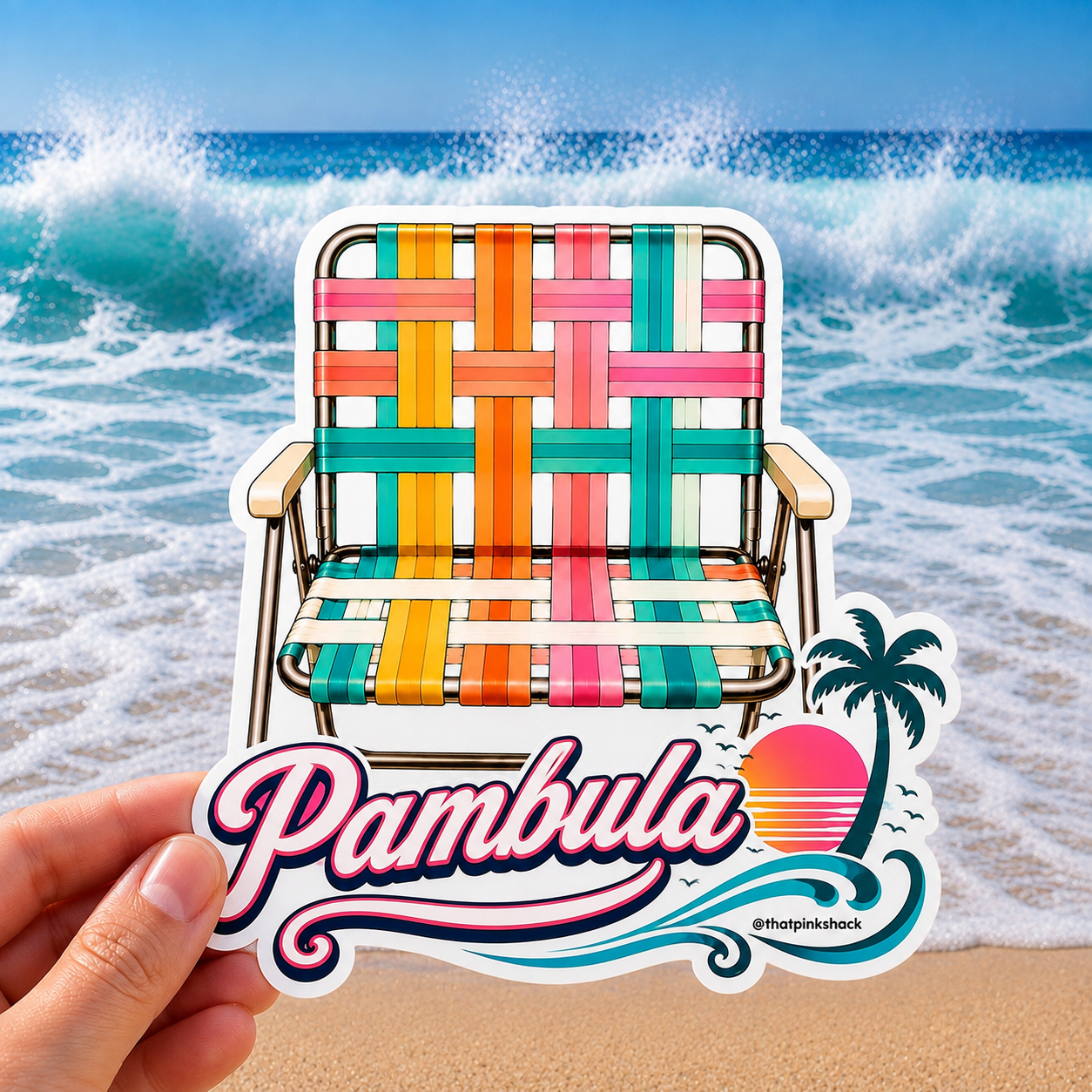 A hand holds a die-cut sticker featuring a colorful woven retro beach chair in front of a beach scene with ocean waves and sand. The sticker includes the text 'Pambula' in large pink script, a palm tree, a sunset
