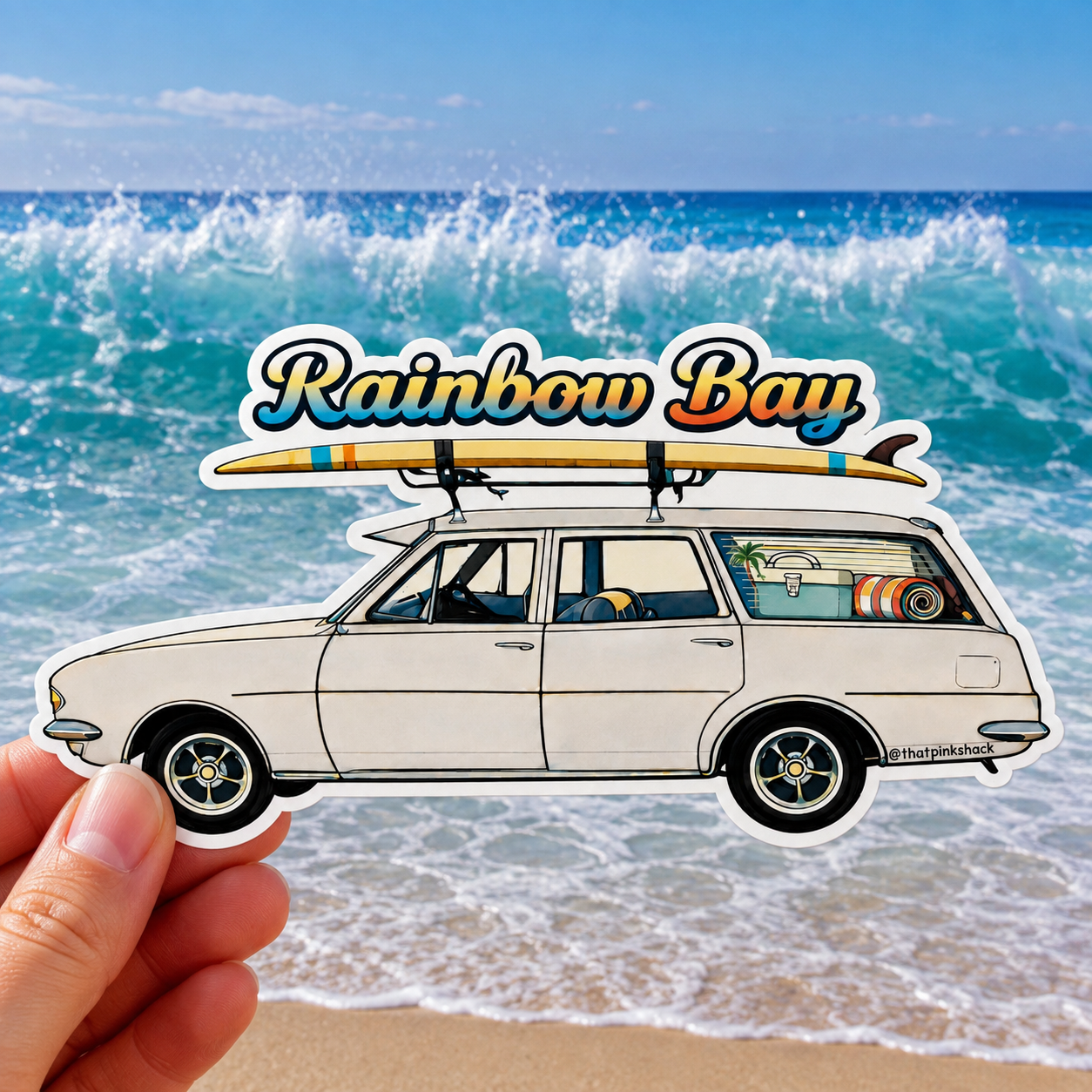 A hand holds a die-cut sticker featuring an off-white vintage station wagon with a surfboard on its roof, set against a beach background with ocean waves and the text 'Rainbow Bay'.