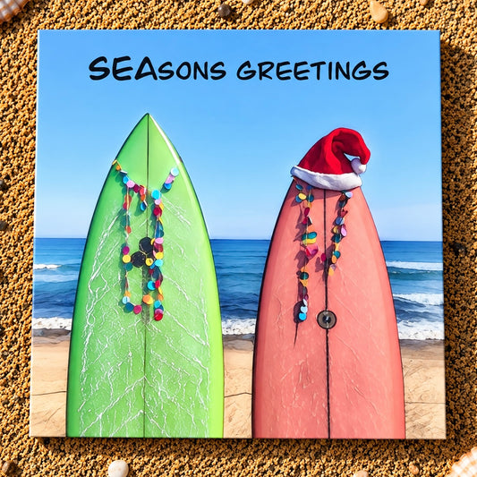 A square printed card with the text 'SEASONS GREETINGS' at the top, showing two surfboards on a beach. One surfboard is bright green with multicolored garlands, and the other is pink with a Santa hat and garlands.