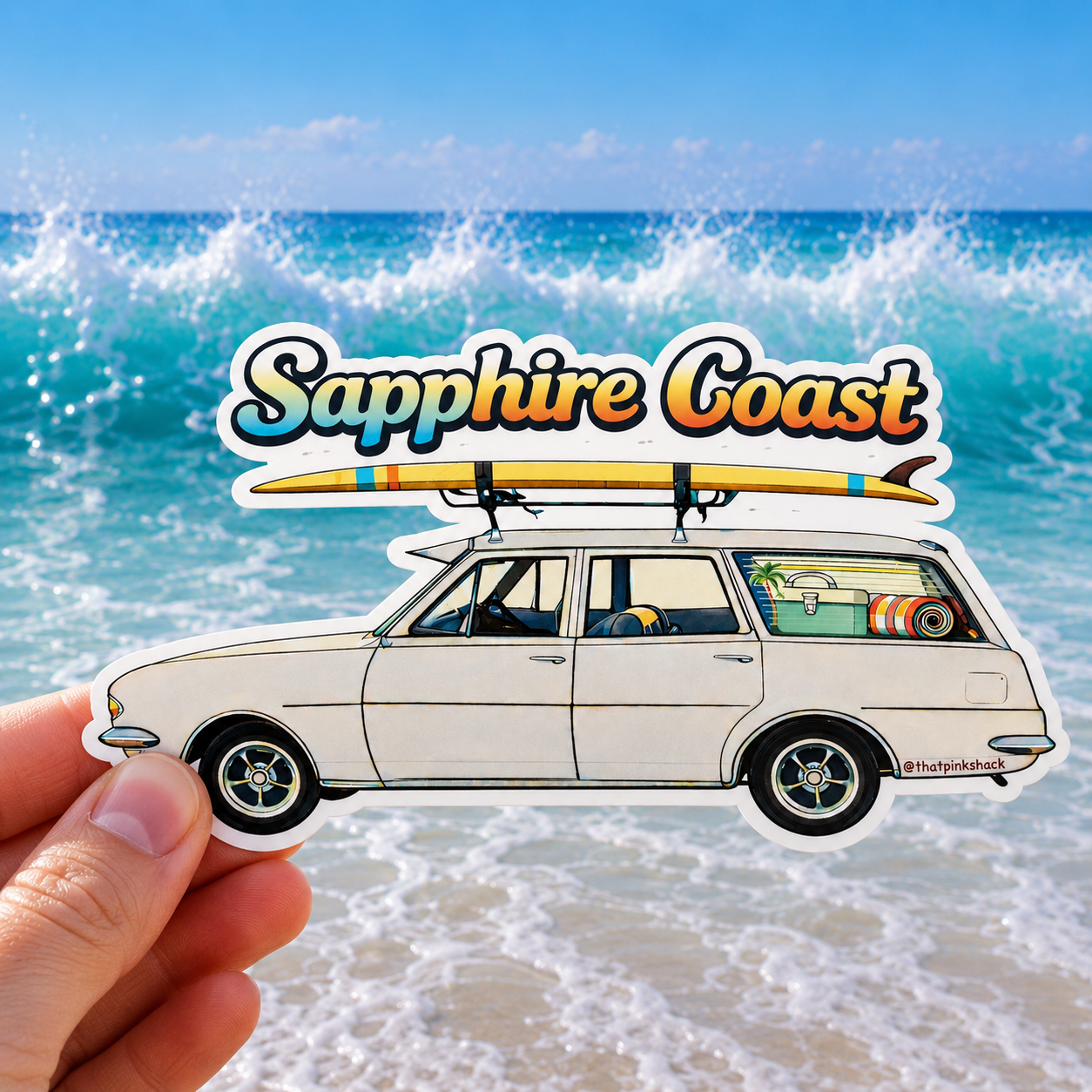 A hand holds a die-cut sticker featuring an illustrated white station wagon with a surfboard on top and the words 'Sapphire Coast' above it, set against a beach and ocean background.