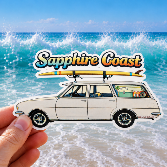 A hand holds a die-cut sticker featuring an illustrated white station wagon with a surfboard on top and the words 'Sapphire Coast' above it, set against a beach and ocean background.