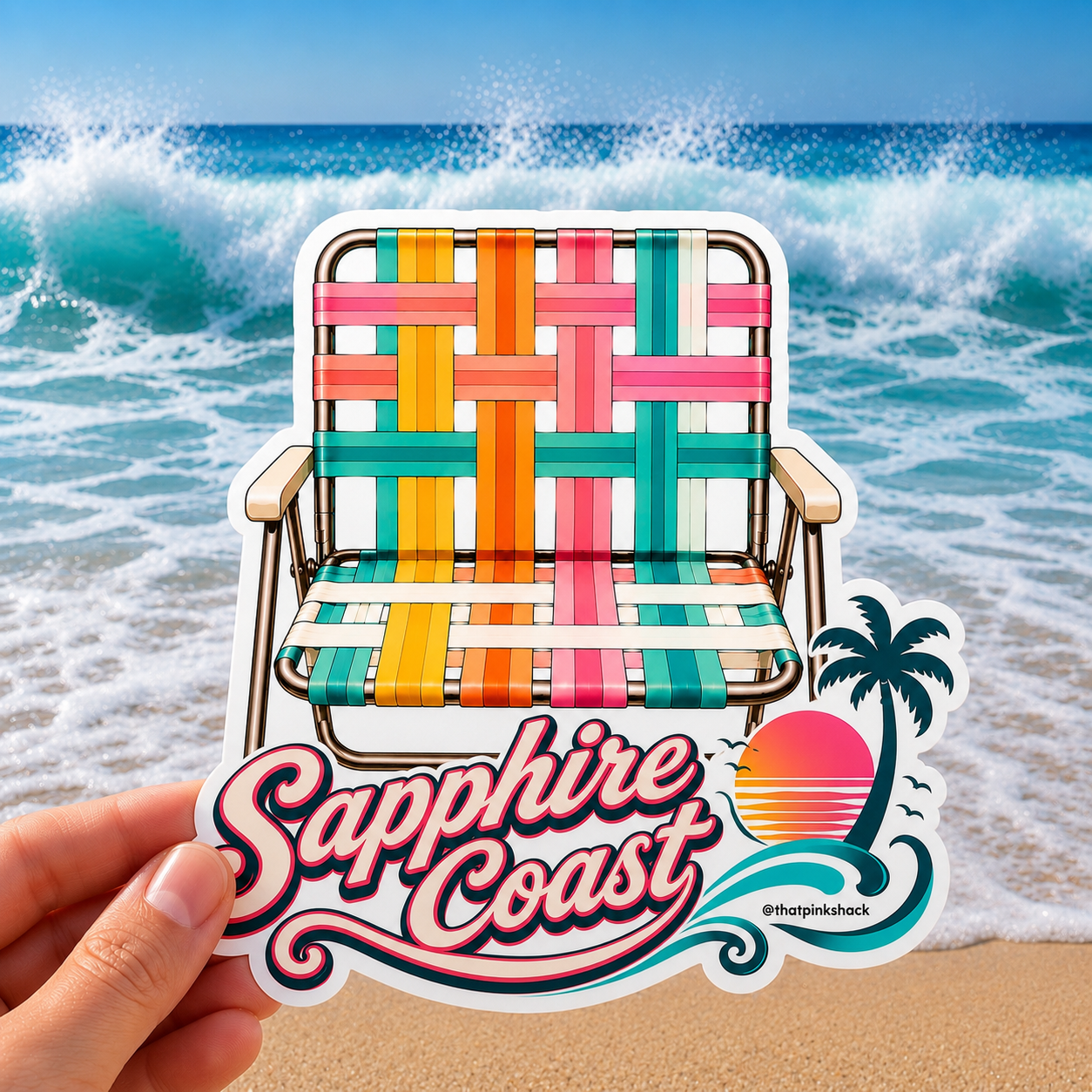 A hand holds a die-cut sticker featuring a colorful retro beach chair and the text 'Sapphire Coast' with a sunset and palm tree, set against a background of ocean waves and sand.