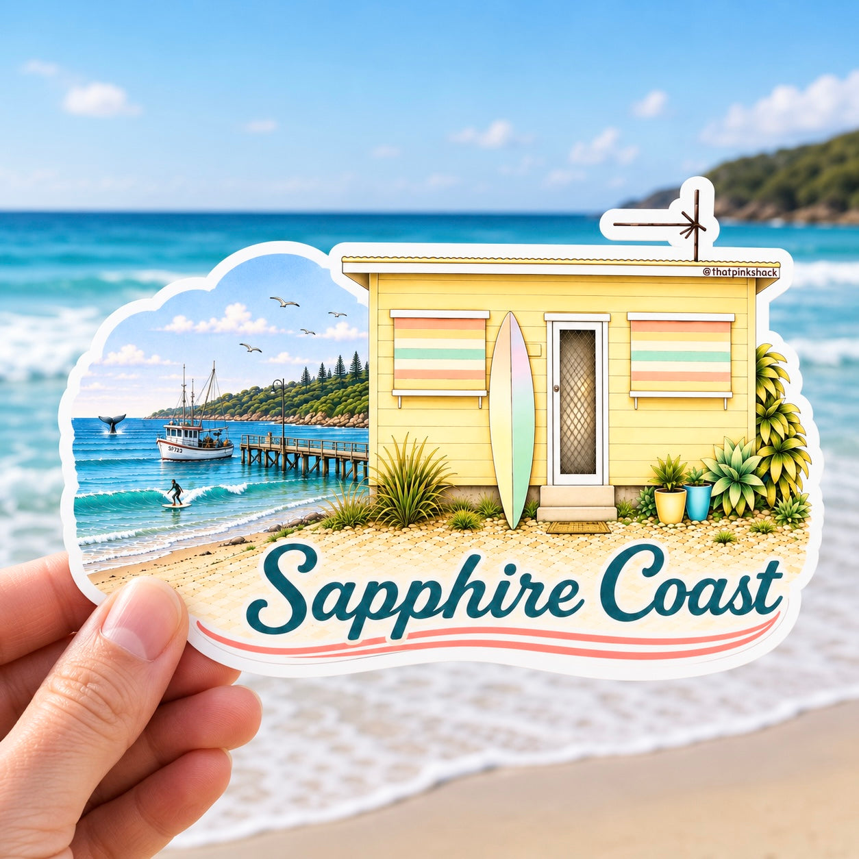 A hand holds a die-cut sticker featuring a yellow beach shack, surfboard, ocean scene, and the text 'Sapphire Coast'. The sticker is shown against a real beach and ocean background.
