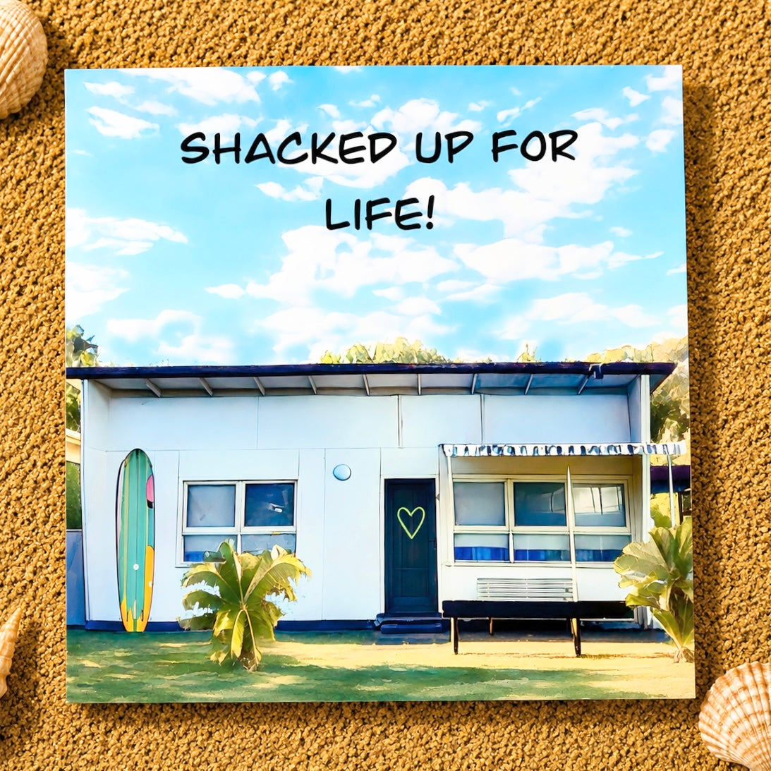 A square greeting card featuring a retro white fibro beach shack under a blue sky with clouds, with the text 'SHACKED UP FOR LIFE!' at the top. The print is styled on a sandy background with shells 