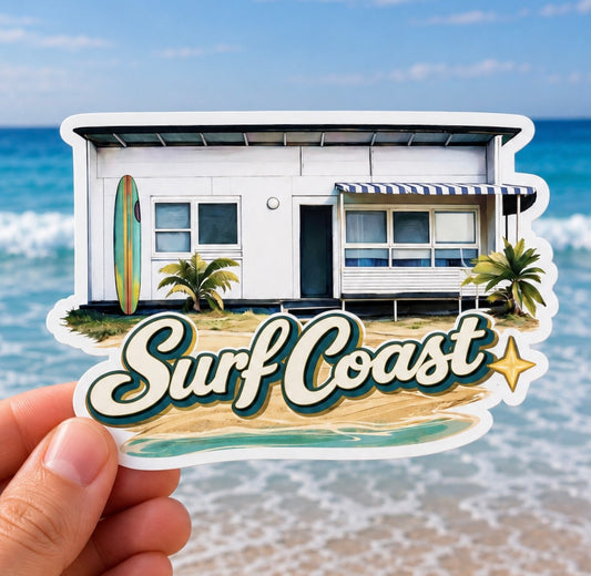 A hand holds a die-cut sticker featuring a retro white beach house with palm trees, a surfboard, and the words “Surf Coast” over sand and ocean water, with a real beach and ocean blurred in the background.