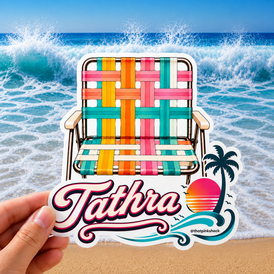 A hand holds a die-cut sticker featuring a colorful woven beach chair and the name 'Tathra' with a palm tree and sunset graphic, set against a background of ocean waves and sand.
