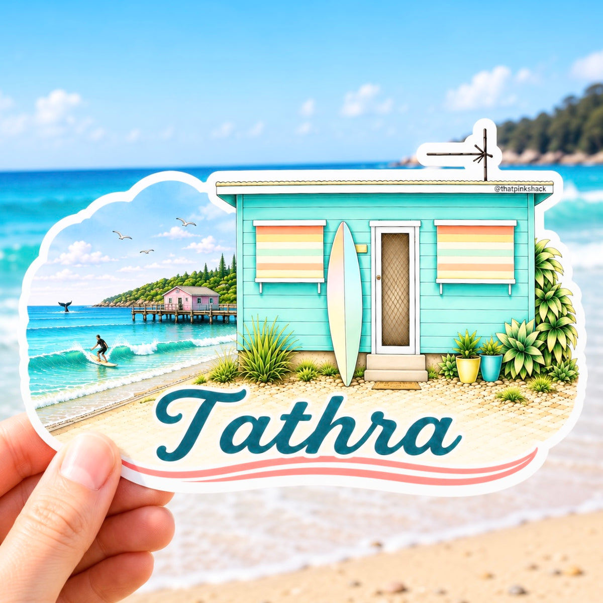 A hand holds a die-cut sticker featuring a colorful beach scene with a turquoise cabana, surfboard, ocean, and the word "Tathra" in large script. The background is a blurred beach and ocean setting.